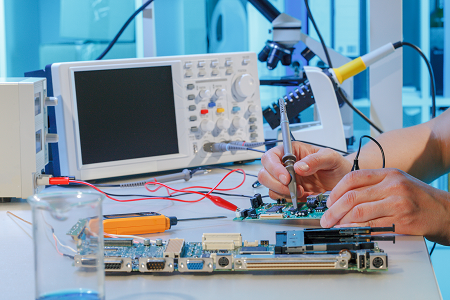 Indoors. Two circuit boards and a couple of electronic testing equipment are on a table. A person holds a test probe over a chip on one of the circuit boards.