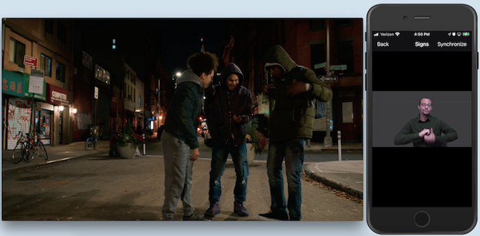 On the left, the image of 3 young men with winter jackets standing next to a street corner, forming a semi-circle. They look at a phone that one of them holds at chest height. On the right, a phone shows the image of an ASL interpreter making a sign.
