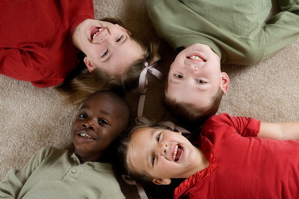 4 children laugh and lay on a carpet. They have their heads together and their bodies pointing in different directions, like forming an X.