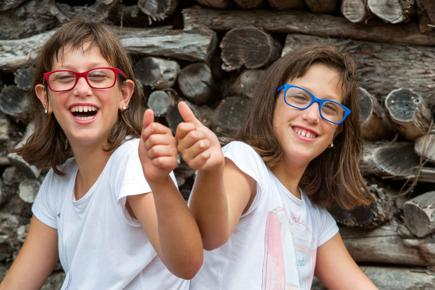 Outdoors. A pair of twin girls with glasses are seated with their backs facing each other. Both girls smile and have turned to look at the front. Each one raises the arm that faces the front and holds the hand at shoulder level. They have their thumb up and the other fingers make a fist.